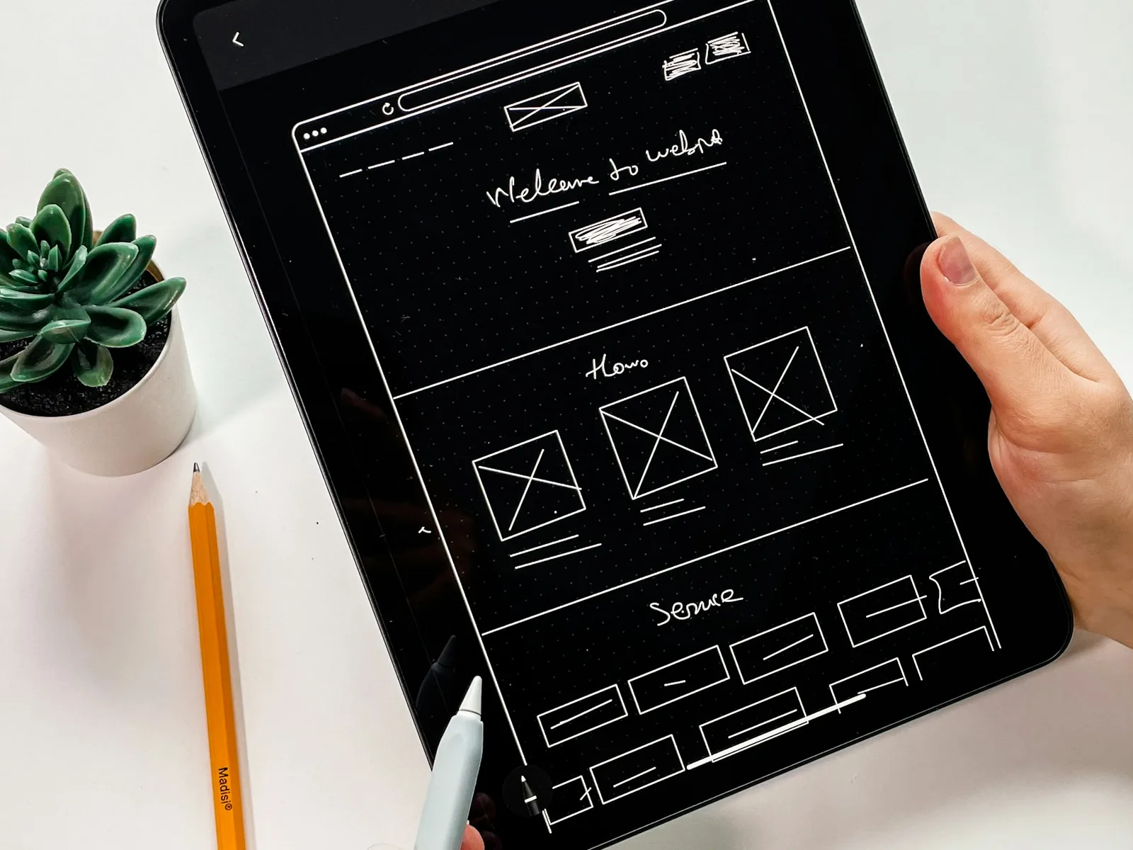 Hände halten ein Tablet mit einem gezeichneten, in Abschnitte gegliederten Seitenentwurf.