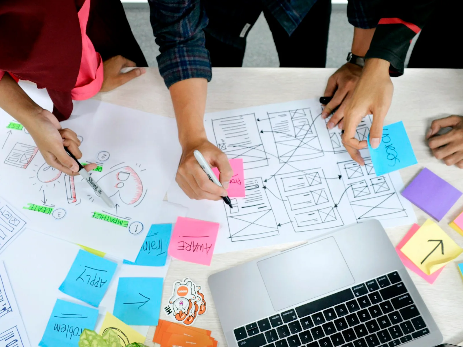 Hände arbeiten gemeinsam an mehreren Website-Wireframes auf einer Tischfläche.