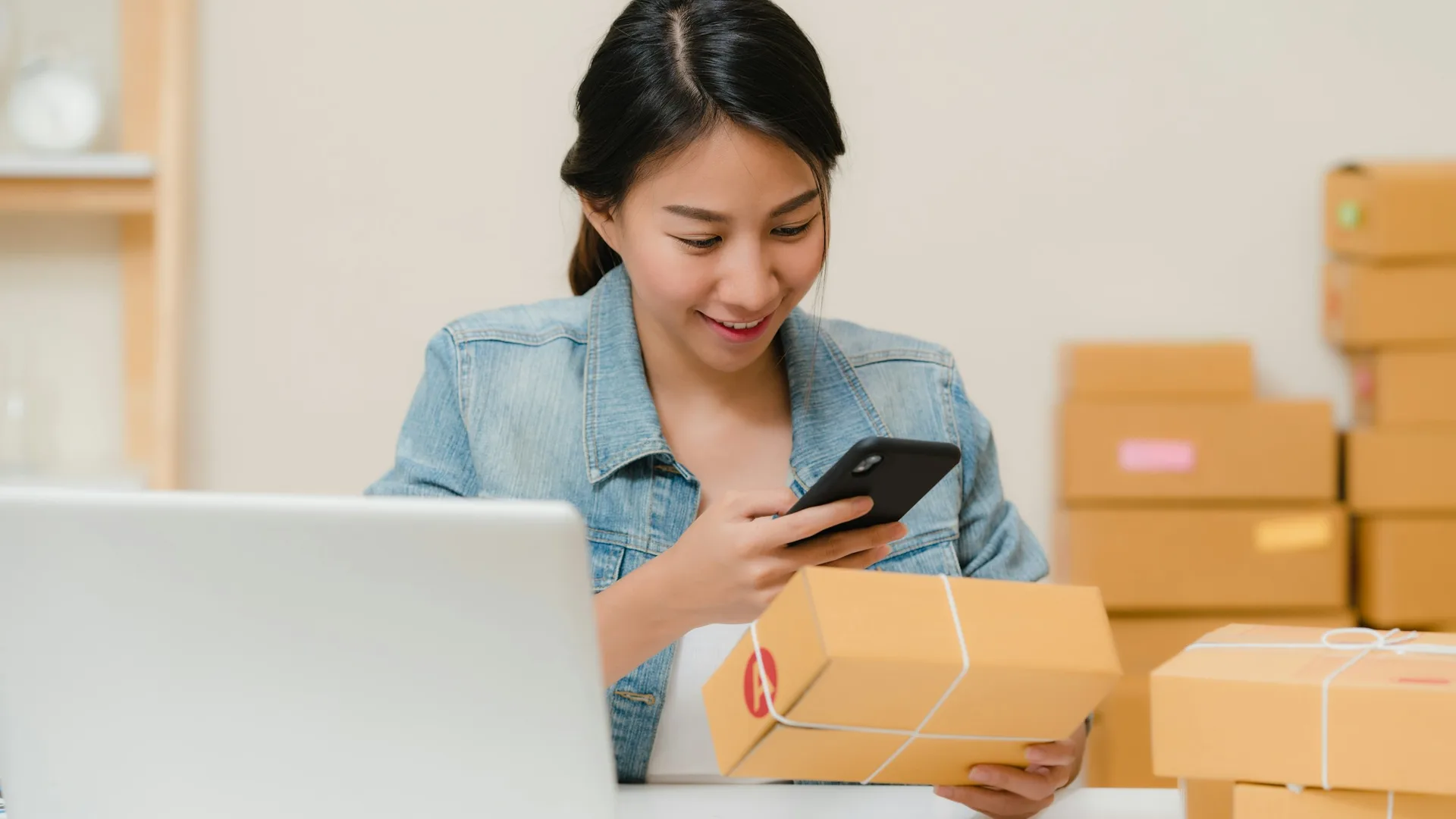Eine Shopbetreiberin prüft ein Paket mit dem Smartphone zwischen Versandkartons.